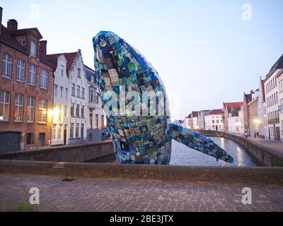 Statua di una balena blu fatta con bottiglie di plastica e altri rifiuti in un fiume a Brugge, Belgio, con lo sfondo di tipici edifici fiamminghi Foto Stock