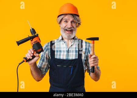 Immagine di un ottimista sorpreso anziano grigio-capelli bearded costruttore di uomo in casco che posa isolato su sfondo parete giallo tenendo trapano e martello. Foto Stock