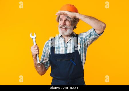 Immagine di positivo ottimista anziani grigio-capelli bearded man builder in casco che posa isolato su parete gialla sfondo tenendo chiave cercando asid Foto Stock