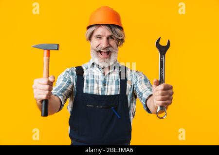 Immagine di allegro anziano anziano grigio-capelli bearded uomo costruttore in casco che posa isolato su parete gialla sfondo tenendo chiave e martello. Foto Stock