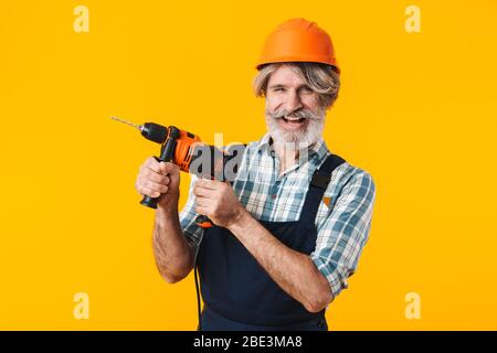 Immagine di felice positivo anziani grigio-capelli bearded uomo costruttore in casco che posa isolato su sfondo giallo parete di tenuta trapano. Foto Stock