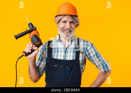 Immagine di felice positivo anziani grigio-capelli bearded uomo costruttore in casco che posa isolato su sfondo giallo parete di tenuta trapano. Foto Stock