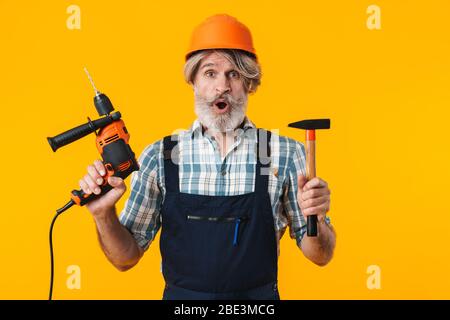 Immagine di un anziano scioccato uomo costruttore barbuto con capelli grigi in posa casco isolato su sfondo parete gialla che tiene trapano e martello. Foto Stock