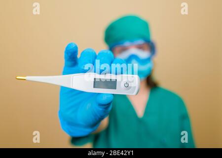 Dottore in tuta verde e guanti blu maschera e occhiali protettivi con termometro in mano su sfondo giallo. Messa a fuoco selettiva. Foto Stock