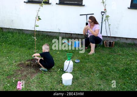 Mamma fotografare figlio annaffiare il nuovo albero di mele piantato nel cortile. Zawady Gmina Rzeczyca Polonia Foto Stock