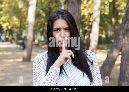 Ritratto di giovane donna indiana felice sorridente, guardando macchina fotografica con i capelli lunghi scuri che tengono il suo indice da labbra isolati alberi verdi parco backgrou Foto Stock