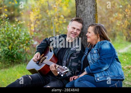 Una coppia matura che passa tempo di qualità insieme e la moglie sta ascoltando suo marito che canta e suona la sua chitarra mentre si trova in un parco della città su un... Foto Stock