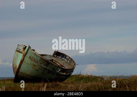 Imbarcazione da pesca abbandonata sull'isola gallese di Anglesey, inclinabile su un lato adagiato sull'erba con un cielo nuvoloso sullo sfondo Foto Stock