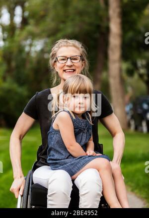 Una mamma paraplegica che tiene la sua bambina in grembo mentre si siede nella sua sedia a rotelle nel suo cortile anteriore su un caldo pomeriggio estivo Foto Stock