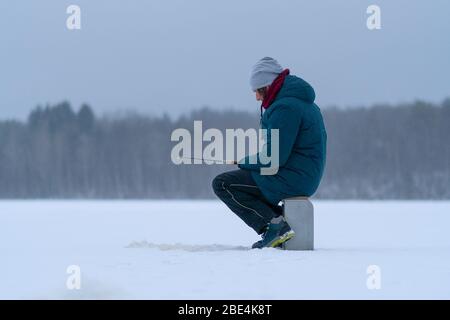 Un uomo su un lago ghiacciato innevato. Vista laterale. Pesca invernale. Canna da pesca nelle mani. Foro di ghiaccio. Neve bianca. Foresta innevata e riva del lago all'orizzonte. Cielo grigio. Foto Stock