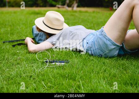 Smartphone con power bank e donna addormentata. Giovane donna dorme su erba verde con cappello che copre il viso mentre dispositivi elettronici portatili in carica. Foto Stock