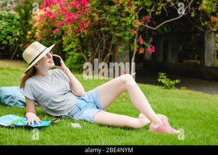 Faccia di giovane donna tenere lo smartphone vicino all'orecchio con l'interruttore sul cavo, indossare cappello e occhiali da sole, a sfondo verde natura da vicino Foto Stock