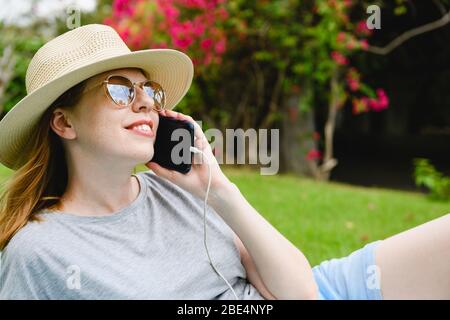 Faccia di giovane donna tenere lo smartphone vicino all'orecchio con l'interruttore sul cavo, indossare cappello e occhiali da sole, a sfondo verde natura da vicino Foto Stock