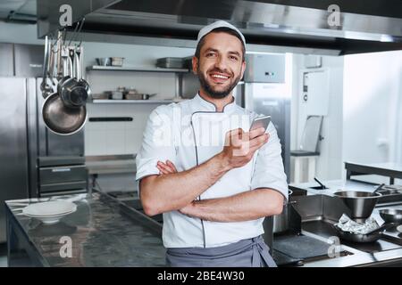 L'uomo barbuto che indossa l'uniforme sta andando cucinare. Guarda la fotocamera con un sorriso, mentre tiene il telefono Foto Stock