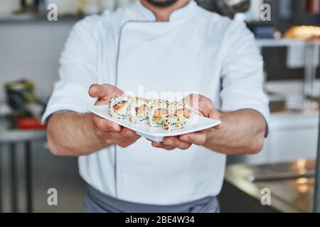 Lo chef giapponese ha preparato sushi al ristorante. Sta in piedi in cucina commerciale e sta tenendo il suo piatto. Immagine ritagliata. Vista frontale Foto Stock