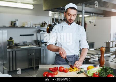 Chef caucasico concentrato in uniforme sta facendo il suo lavoro. Trita le verdure per il suo piatto caratteristico. Concetto di cucina Foto Stock