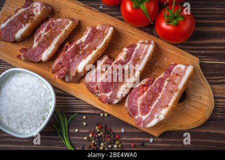Fette crude di pancetta di maiale su un tagliere, su un tavolo rurale in legno. Pancia di maiale con verdure. Foto Stock