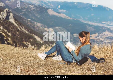 Giovane bella ragazza viaggia da sola in montagna in primavera o autunno, siede sul bordo della montagna e libro di lettura e gode di natura, rocce e foreste verdi, vista del paesaggio. Uno zaino dietro e abbigliamento sportivo, un thermos con una bevanda calda o tè, libertà e leggerezza. Vista posteriore della donna viaggiatore in CAP seduto solo sulla cima della montagna e guardando il bellissimo paesaggio estivo e vista mare blu. Bere tè o caffè Foto Stock