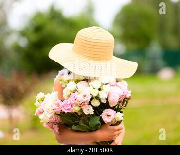 La ragazza tiene un grande bouquet di peonie rosa e bianca davanti a lei. Un bambino in un cappello di paglia con ampio bordo. Gode del profumo dei fiori. Giornata estiva soleggiata. Foto Stock