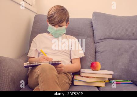 Bambino ragazzo che studia online a casa con libri, indossare maschera facciale protettiva, seduta su elegante divano grigio vicino alla finestra. E-learning, concetto di soggiorno a casa Foto Stock