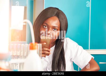 Giovane donna africana sorridente scienziato, laureato o studente medico con il telefono cellulare guarda la fotocamera. La donna sorride mentre usa un dispositivo digitale Foto Stock