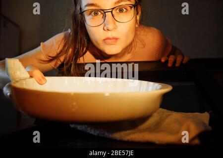 Vista dal forno. La ragazza prende una padella di vetro dal forno. La persona è illuminata dalla luce della lampada del forno. Foto Stock