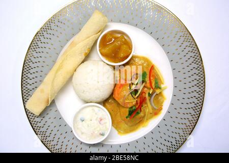 Una cena indiana pronta per servire con tikka masala di pollo, riso, chutney, raita e un pane cappati, su un piatto grigio ornato e panno da tavolo bianco. Foto Stock