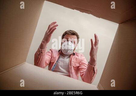 Uomo stupito in maschera medica disboxing all'interno di vedere una scatola consegnato. Concetto di cautela e protezione durante un focolaio di virus. Foto Stock