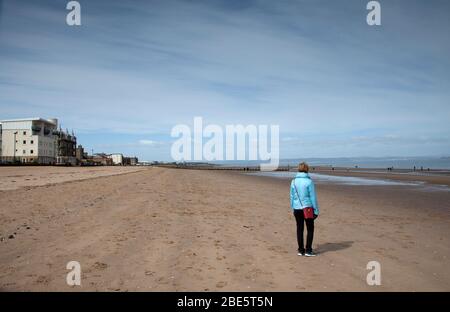 Portobello, Edimburgo, Scozia, Regno Unito. 12 aprile 2020. Frizzante con occasionale sole al mare. Eccezionalmente tranquilla sulla Promenade e sulla spiaggia nel primo pomeriggio della Domenica di Pasqua a causa del pubblico che soggiorna a casa a causa del Coronavirus Lockdown, Donna in primo piano sulla spiaggia di sabbia dà scala a quanto vuota la spiaggia è. Foto Stock