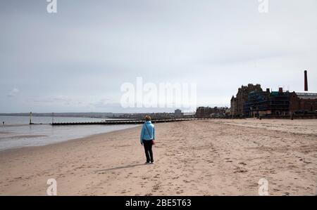 Portobello, Edimburgo, Scozia, Regno Unito. 12 aprile 2020. Frizzante con occasionale sole al mare. Eccezionalmente tranquilla sulla Promenade e sulla spiaggia nel primo pomeriggio della Domenica di Pasqua a causa del pubblico che soggiorna a casa a causa del Coronavirus Lockdown, Donna in primo piano sulla spiaggia di sabbia dà scala a quanto vuota la spiaggia è. Foto Stock