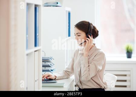 Vista laterale ritratto di donna d'affari sorridente che parla al microfono mentre lavora con il laptop all'interno dell'ufficio, spazio per copiare Foto Stock