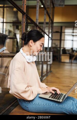 Immagine di una giovane bella donna asiatica che lavora su un notebook mentre si siede a pavimento in ufficio Foto Stock