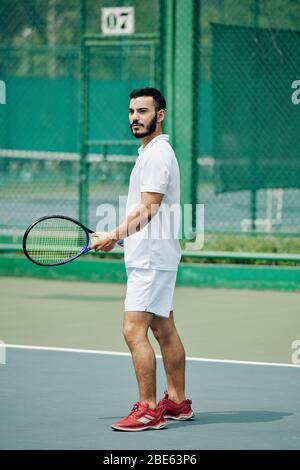 Bel giovane ispanico in abbigliamento sportivo bianco giocando a tennis all'aperto Foto Stock
