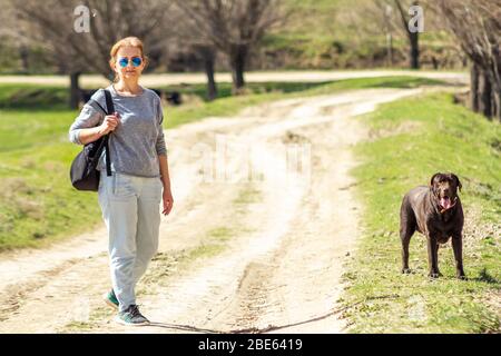 Donna in occhiali da sole che rimane su una strada, con cane cioccolato Labrador Retriever. Foto Stock