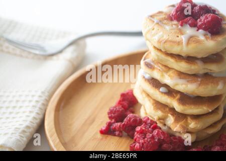 Frittelle soffici con latte condensato e lamponi su un piatto di legno con tovagliolo da cucina e forchetta Foto Stock