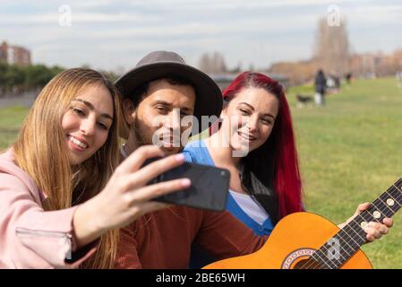 Un ragazzo latino con chitarra si posa mentre si scatta una foto con due ragazze caucasiche nel parco cittadino Foto Stock