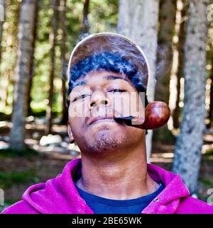 felice giovane uomo fuma con piacere il tabacco nella sua pipa tra dolci nuotate e nuvole di fumo Foto Stock