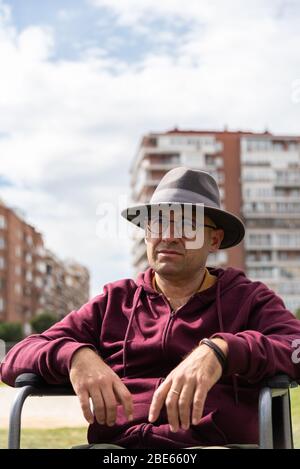 Uomo caucasico con occhiali e cappello depressivo e triste in una sedia a rotelle solitaria in un parco con il cielo e alcuni edifici sullo sfondo con lo spazio Foto Stock