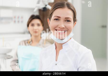 Un primo piano di una dottoressa sorridente alla telecamera, con il paziente in attesa di essere controllato in background. Foto Stock