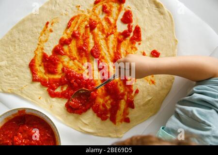 Il ragazzino finisce di mettere la salsa di pomodoro con un cucchiaio su una pizza Foto Stock