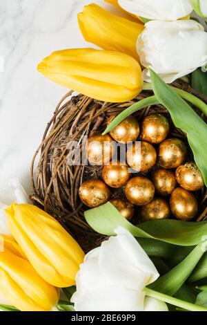 Bel bouquet di tulipani gialli e bianchi su tavolo in marmo. Quaglia d'oro dipinta uova in nido rustico. Felice Pasqua concetto. Vista dall'alto. Spazio di copia Foto Stock