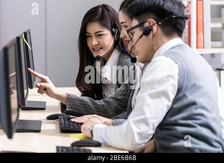 Operatore asiatico di fiducia e cordiale per giovani adulti con cuffie e auricolari che lavorano in un call center con il suo team di colleghi che lavora come customer servic Foto Stock