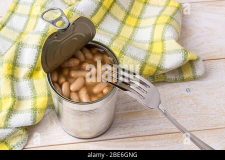 Fagioli in scatola in una lattina aperta con linguetta di trazione e una forchetta su un tavolo di legno. Fonte di proteine vegetali e ingrediente per cibo vegetariano. Foto Stock