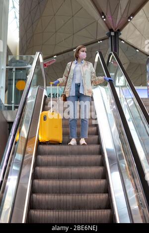 Donna con bagaglio giallo si trova sulla scala mobile presso il terminal dell'aeroporto quasi vuoto a causa delle restrizioni di viaggio per l'epidemia di coronavirus pandemico/Covid-19. Flig Foto Stock