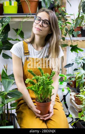 Donna giardiniere in occhiali che indossano tute, tenendo un felce in vaso di plastica, piante casalinghe in vaso sullo sfondo. Giardinaggio domestico, amore delle piante domestiche, libero Foto Stock