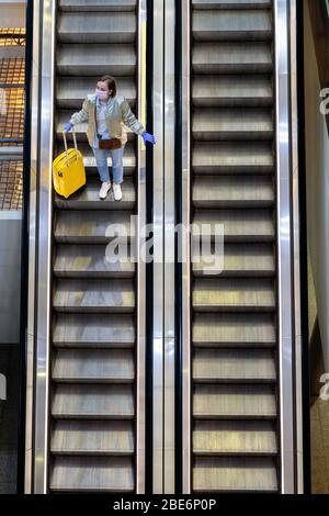 Donna con bagaglio giallo si trova sulla scala mobile presso il terminal dell'aeroporto quasi vuoto a causa delle restrizioni di viaggio per l'epidemia di coronavirus pandemico/Covid-19. Flig Foto Stock