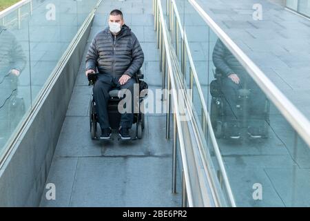 Uomo disabile su sedia a rotelle che indossa una maschera medica per la protezione del coronavirus. Foto Stock