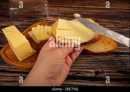 Qualcuno spalma il burro su pane tostato di grano fresco con un coltello e un piatto di burro di legno con un pezzo di burro e fette di formaggio su uno sfondo di legno. Foto Stock