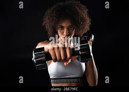 Immagine di fidata sportivo afro-americana facendo esercizio con manubri isolati su sfondo nero Foto Stock