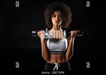 Immagine di fidata sportivo afro-americana facendo esercizio con manubri isolati su sfondo nero Foto Stock